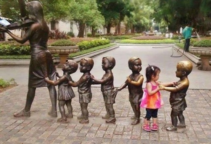 A little girl wearing a pink top stands at the end of a line of bronze children holding each other’s shoulders behind a woman playing violin in a public square.