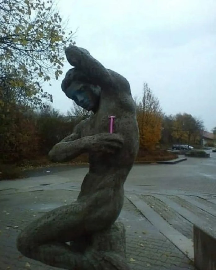 A grey stone statue of a muscular male figure with his arm raised has a real pink razor placed in his armpit, creating a humorous modern twist.