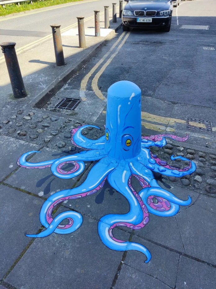 A bright blue bollard painted as a whimsical octopus with pink tentacles extending across the sidewalk, created by Lumen Street Theatre in Limerick, Ireland for one of their scavenger hunts.