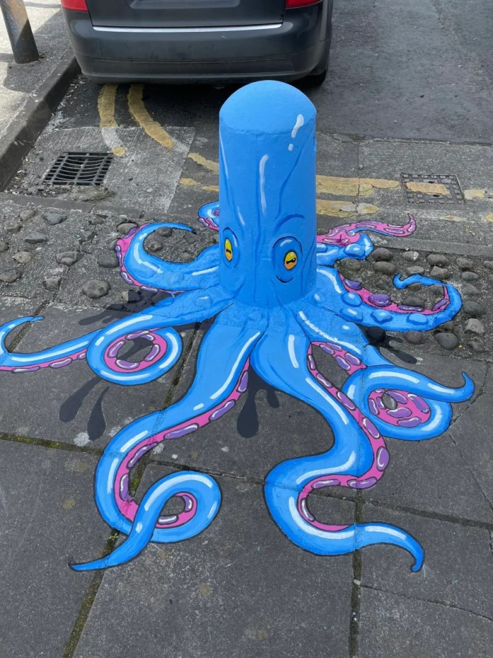 A bright blue bollard painted as a whimsical octopus with pink tentacles extending across the sidewalk, created by Lumen Street Theatre in Limerick, Ireland for one of their scavenger hunts.