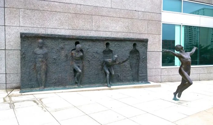 A series of bronze figures appear to push out from a textured wall, with one human form breaking fully free in a triumphant pose on a city sidewalk in Philadelphia.