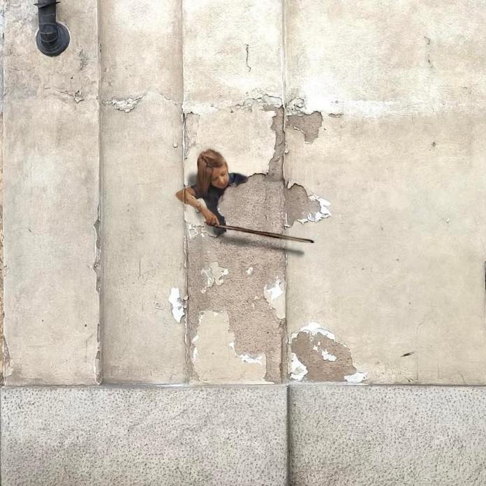 A mural on a cracked wall shows a girl holding a violin bow as if playing an invisible double bass, with the peeling plaster acting as the instrument strings, painted by Golsa Golchini in Milan.