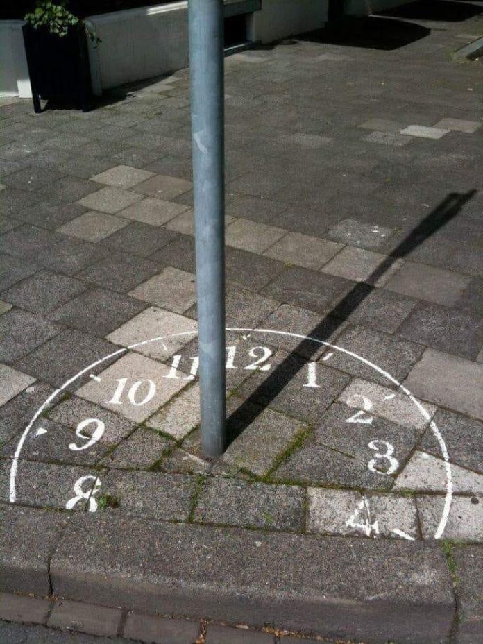 A sidewalk painted like a clock face uses a vertical metal pole’s shadow to indicate the hour, creating a functional street sundial surrounded by paving stones.