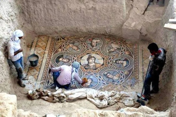 Large circular Roman mosaic from Zeugma featuring eight portrait medallions surrounding a central youthful figure, unearthed in a stone chamber with archaeologists cleaning the tiles.