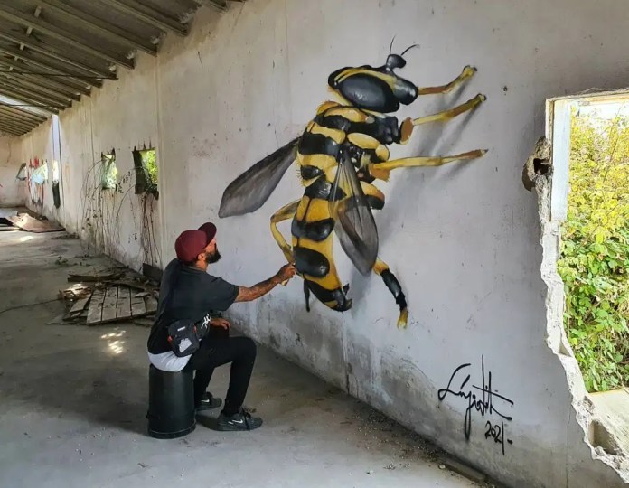 A mural of a hyperrealistic bee, much larger than life-size, appears to land on a wall as the artist reaches toward it with his paintbrush.