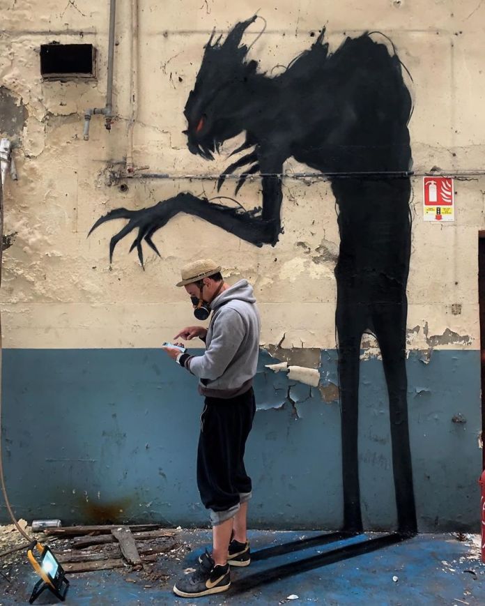 A tall, black shadow figure with long arms and red eyes is painted behind a man standing in an abandoned space, making it appear as his sinister shadow.