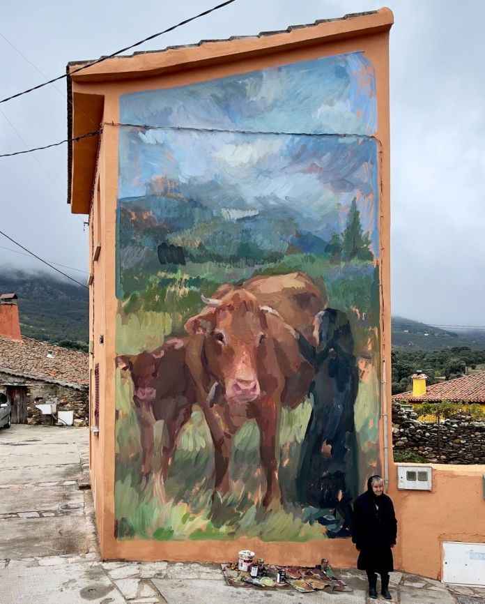 Mural with expressive brushwork depicting three cows standing on grassy hills with a mountainous background. A real elderly woman stands at the base for scale.