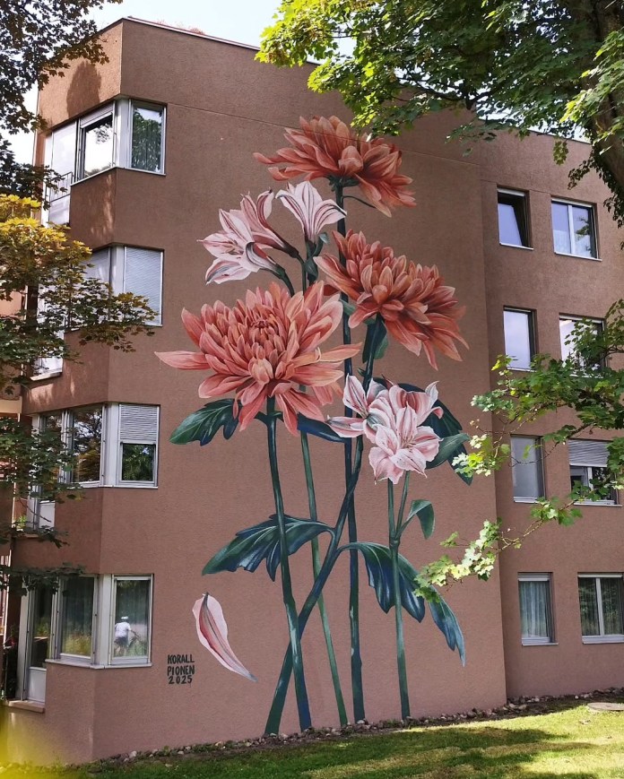 Wall mural of oversized coral-pink and white peony flowers with green stems and leaves, covering multiple stories of a brown apartment building.