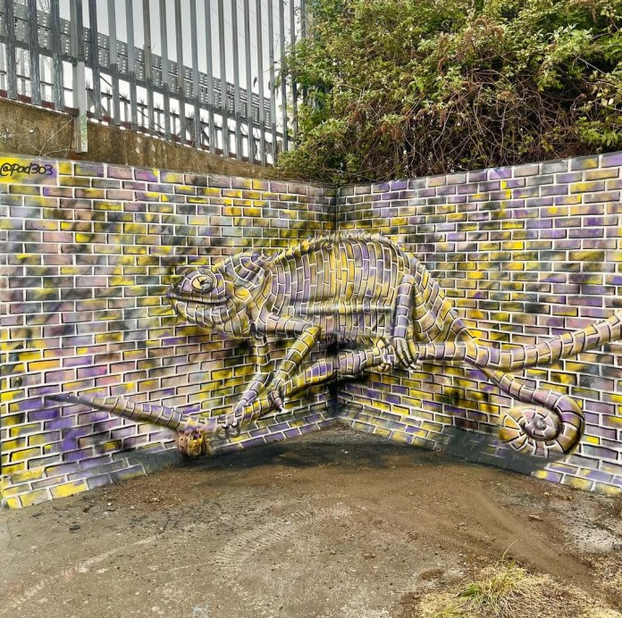 Trompe-l'œil mural of a chameleon painted in yellow and purple tones on a brick corner wall, blending into the background. Created by Paddy Watts.