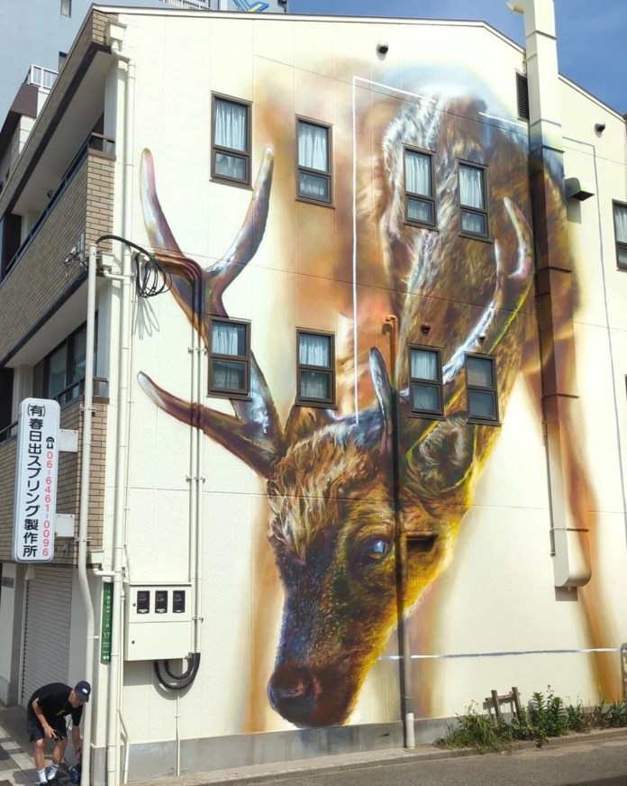 Large deer mural in golden and brown tones painted on a multi-story white building, with antlers wrapping around windows. Located in Osaka, Japan.