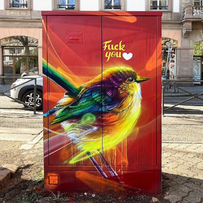 Bright mural of a rainbow-colored bird painted on a red utility box with graffiti text reading “Fck you ♥.” Street art by DAN23 in Strasbourg, France.