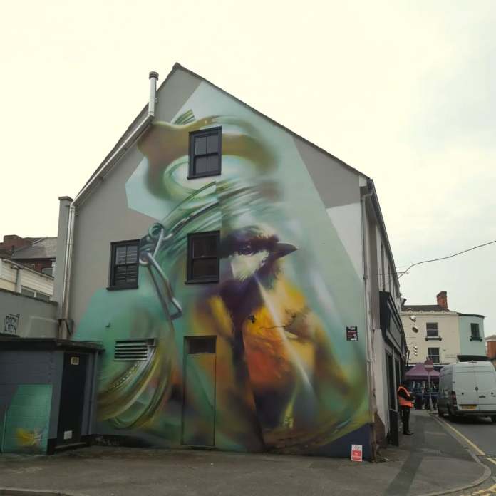 Mural of a songbird appearing trapped inside a glass jar, painted on the side of a house in Swindon, England. The jar’s distorted glass and metal clasp are depicted with realistic lighting and transparency effects, enhancing the mural’s emotional weight.