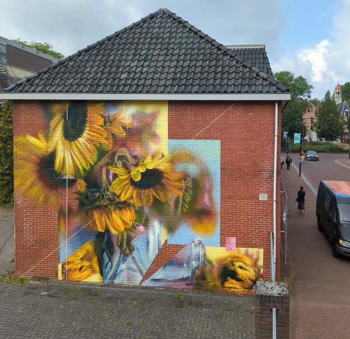 Mural of multiple sunflowers in various stages of bloom and decay, painted on a red brick wall in Assen, Netherlands. The flowers are realistically detailed, with some petals vibrant and others wilting, symbolizing natural cycles of life.