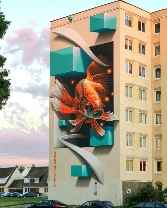 A massive 3D mural of a goldfish appearing to swim through and around turquoise cubes on the side of a residential building in Calais, France. Created by Sweo and Nikita.
