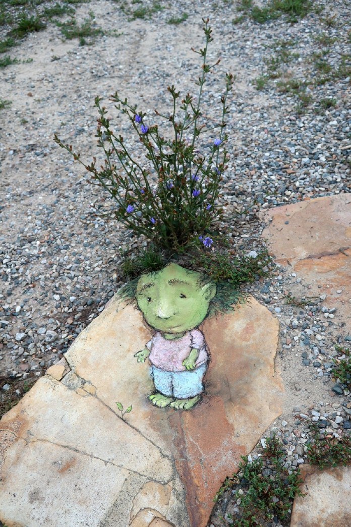 Chalk drawing on a large stone depicting a small green-skinned figure in a pink shirt and blue pants, positioned so grass and purple flowers sprouting from a crack behind the stone look like the figure’s hair.