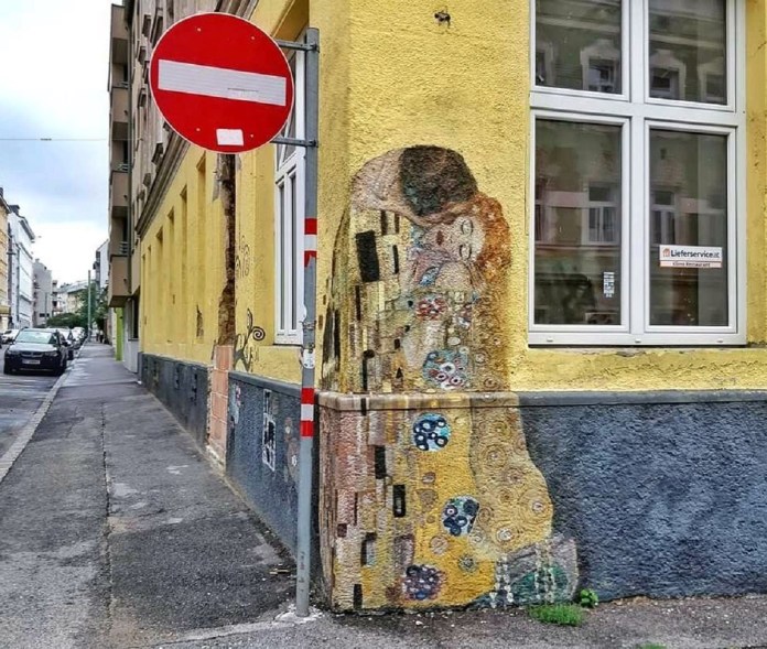 Corner mural reproducing Gustav Klimt’s “The Kiss” on a yellow wall, positioned so a red-and-white no-entry street sign partly covers the embracing couple.