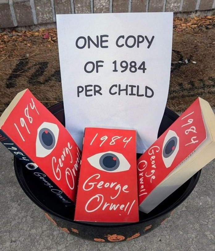 Plastic bowl on pavement containing several red-cover copies of George Orwell’s 1984, with a sign reading “One copy of 1984 per child” propped behind them.