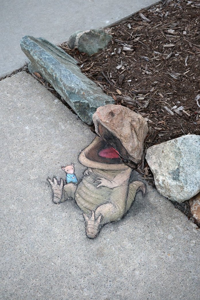 Chalk art on a sidewalk where a rock is used as the creature's head, forming a dinosaur-like body with a smiling face and a small cartoon pig beside it.