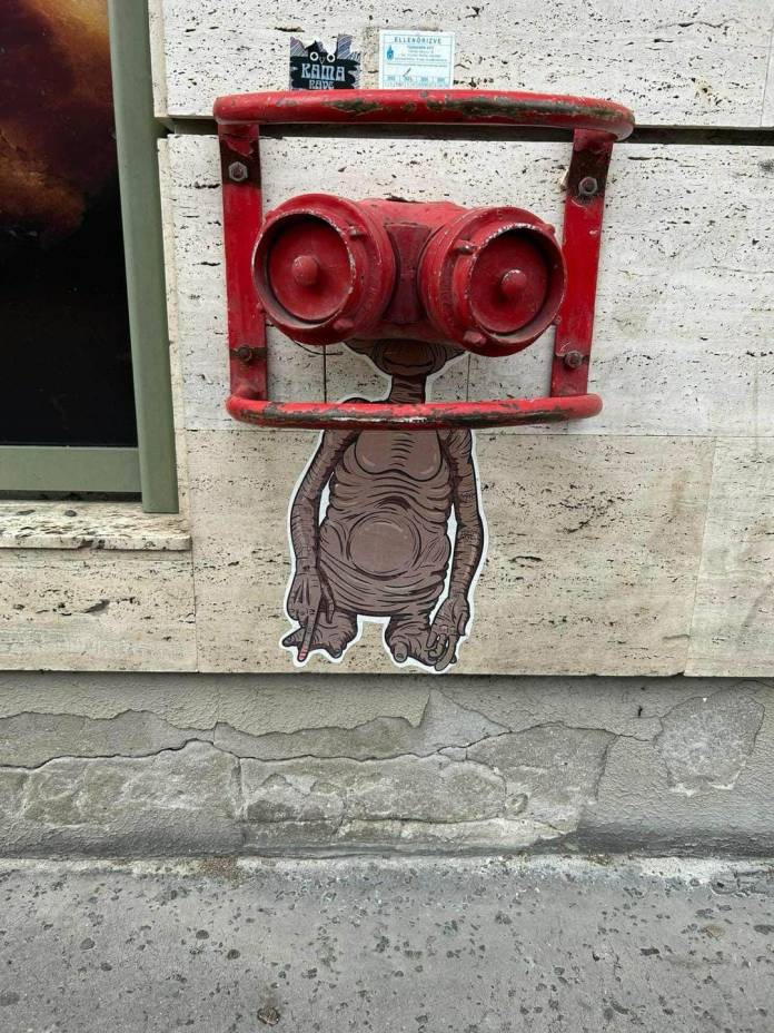 A red metal fire hydrant fixture on a wall combined with a pasted illustration of E.T.'s body to create the illusion of his full figure using the fixture as his head.