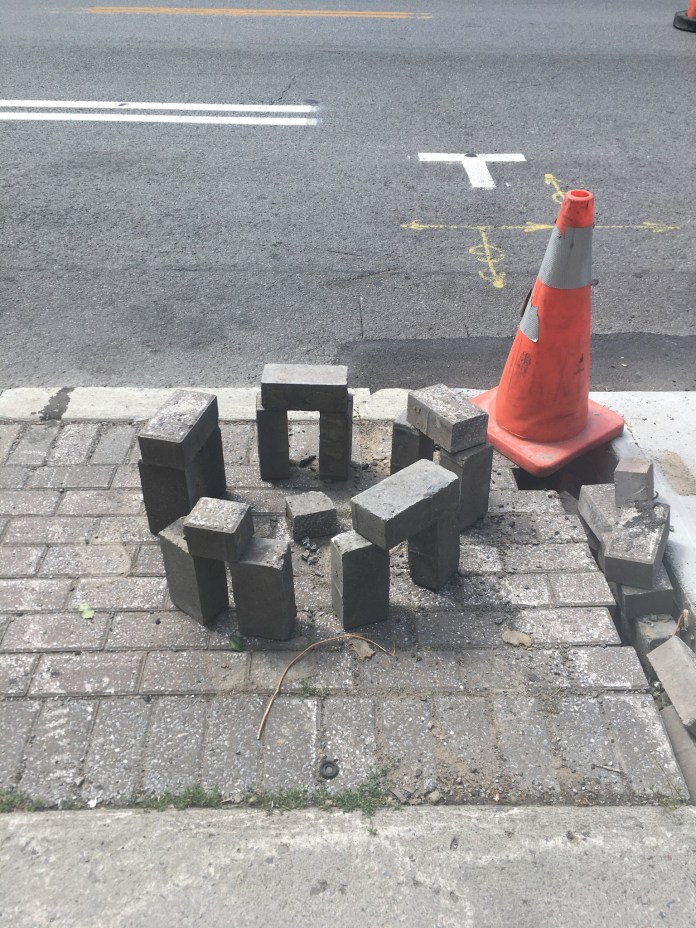 A playful arrangement of sidewalk bricks mimicking Stonehenge, placed around a small open pit next to a traffic cone on a city sidewalk.