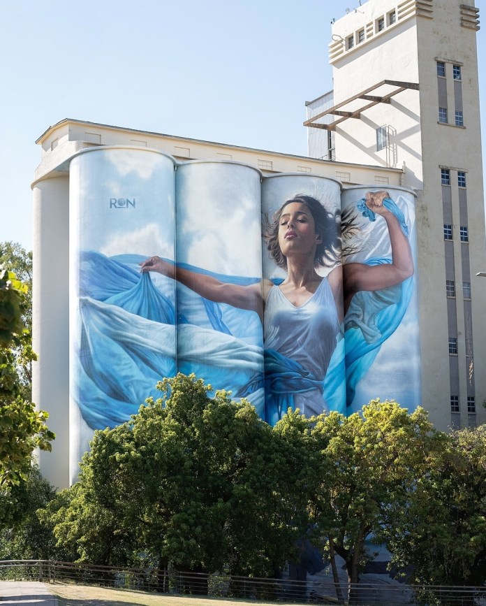 Gigantic mural by Martín Ron in Rosario, Argentina, showing a woman in a flowing blue dress standing across four cylindrical silos, with eyes closed and arms stretched upward, fabric flowing around her in a wind-like motion.