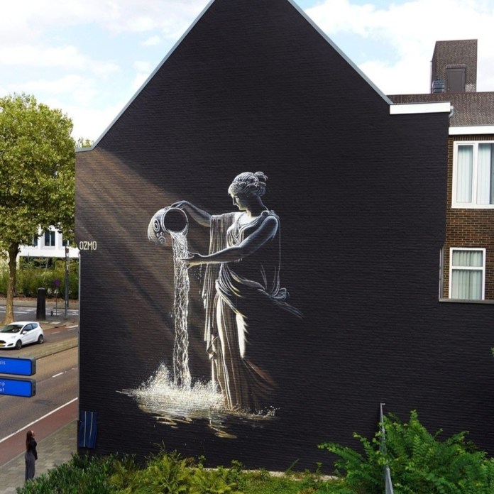 Mural by Ozmo in Heerlen, Netherlands, on a black wall showing a statue-like woman pouring water from a jug, painted in shades of grey and white with realistic water splash and shadow effects.