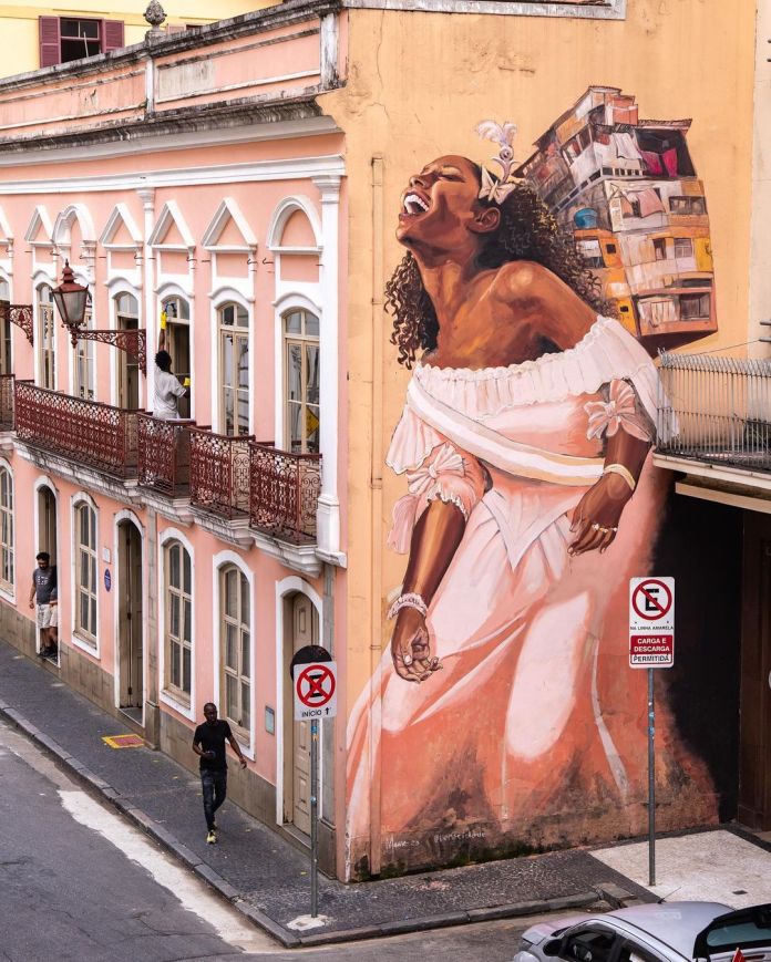 Mural by Veracidade in São Paulo, Brazil, showing a woman in a white dress with curly hair and a headpiece, laughing joyfully with a model of a favela on her back, integrated into the architecture of a pink building.