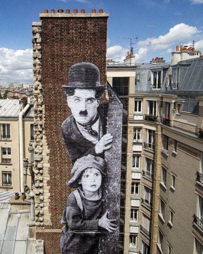 Mural by JR in Paris, France, showing black-and-white photographic wheatpaste of Charlie Chaplin and a child peeking from behind a wall, placed on a tall brick chimney between apartment buildings.
