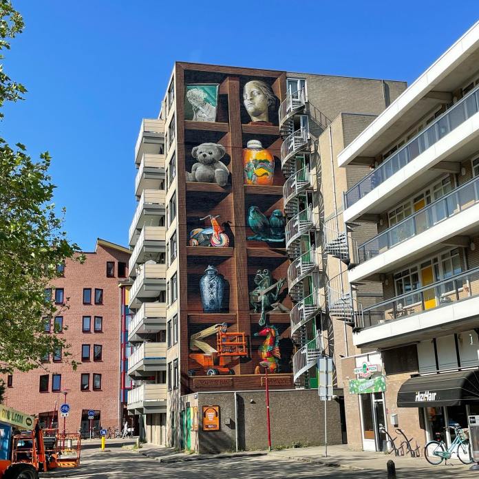 Mural by Jan Is De Man in Nieuwegein, Netherlands, showing a large-scale 3D illusion of a wooden display shelf on the side of an apartment building, filled with realistic painted objects including statues, toys, and vases.