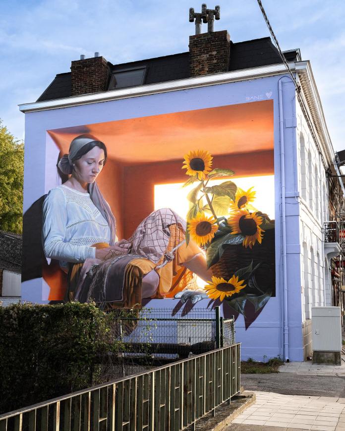 Mural by Bane Florin in Mons, Belgium, depicting a girl with a headscarf sitting in a glowing window, mending a fishing net beside large sunflowers, bathed in golden light.