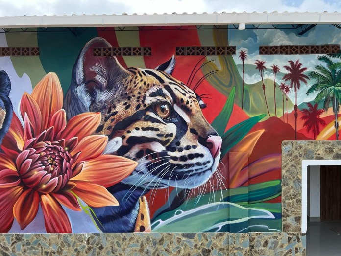 Detailed mural of an ocelot's face with sharp fur textures and golden eyes, surrounded by tropical flowers and palm trees against a vibrant jungle backdrop, painted on a building wall in Barcelona, Colombia.