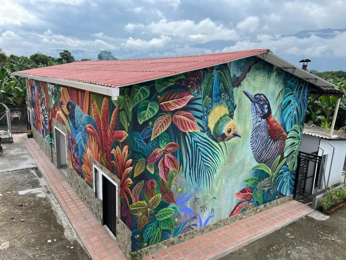 Wide-angle view of a building mural showing two tropical birds, one perched and one flying, surrounded by multicolored leaves and lush vegetation, creating a deep jungle illusion.