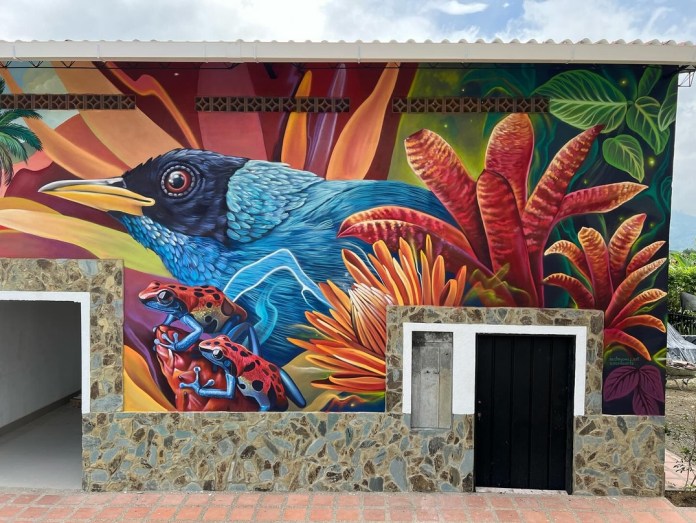 Mural segment showing two vividly colored poison dart frogs below a blue-feathered bird, surrounded by fiery red and yellow leaves, painted on a Colombian building facade.