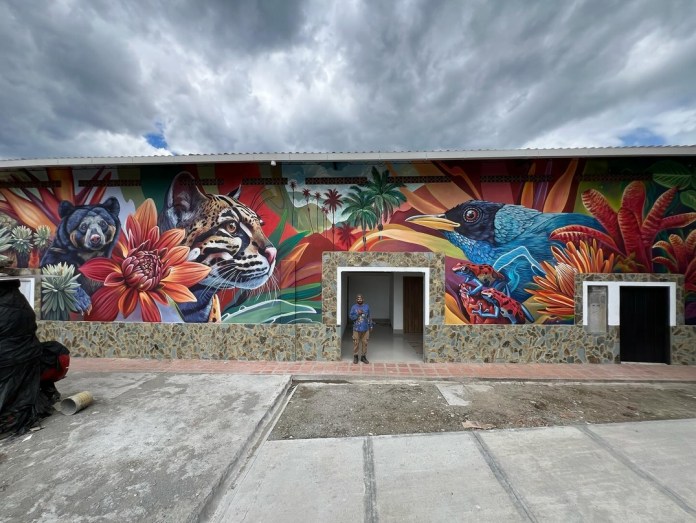 Panoramic view of a full-building mural with an artist standing in front, showcasing an ocelot, bear, tropical birds, frogs, and flowers painted across the entire facade in Barcelona, Colombia.