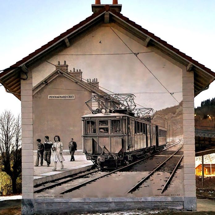 A mural by NESSE in Le Crey-Susville, France, depicting a detailed, vintage-style train station scene in sepia tones with passengers waiting and an electric train pulling in.