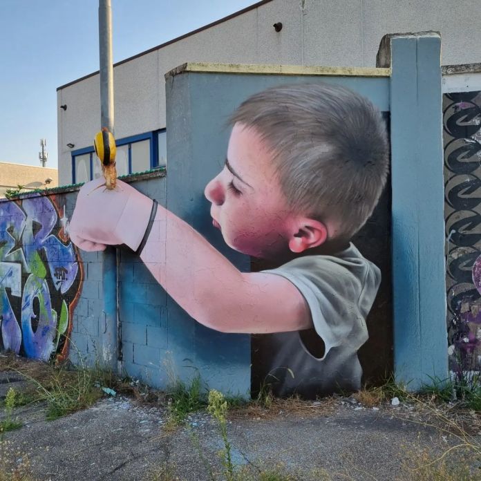 A mural by CHEONE in Italy showing a realistic boy holding a physical striped bollard, with the painted figure integrated seamlessly into the wall’s corner for a playful 3D effect.