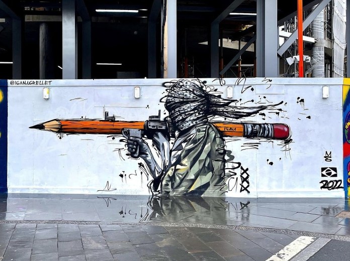 A mural by IGANA in London, UK, of a figure with a headscarf aiming a giant pencil like a bazooka, blending graffiti and political commentary through expressive brushstrokes and contrast.