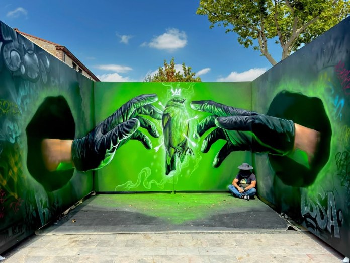 A vivid 3D graffiti mural by Braga last1 at FESTIVAL ULTRA VIOLET in Thuir, France, showing two large realistic hands in black gloves reaching toward a bright green bird pierced by multiple arrows, with a small white crown floating above it. The artwork spans three walls, creating a strong illusion of depth. A person sitting in the corner highlights the mural's large scale.
