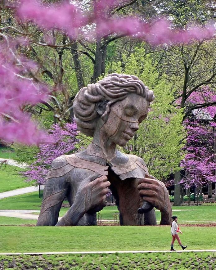 Large outdoor sculpture of a wooden woman with closed eyes and hands opening her chest, placed in a blooming park with green and pink foliage in Lisle, Illinois, USA.
