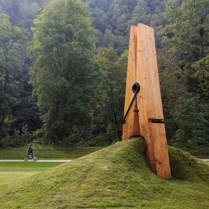 Oversized wooden clothespin sculpture installed in a grassy field, visually pinching the earth to create the illusion of tension in the landscape.