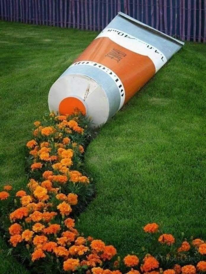 Outdoor installation of a giant paint tube on a green lawn with bright orange flowers planted in a curved line, giving the illusion that the flowers are being squeezed out like paint.