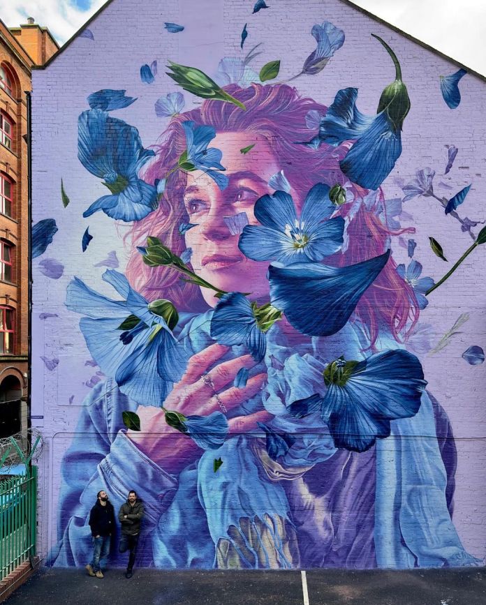 Mural of a woman with curly red hair wrapped in a blue scarf, surrounded by swirling flax flowers in various shades of blue, painted on a lavender wall in Belfast, Northern Ireland by Studio Giftig.