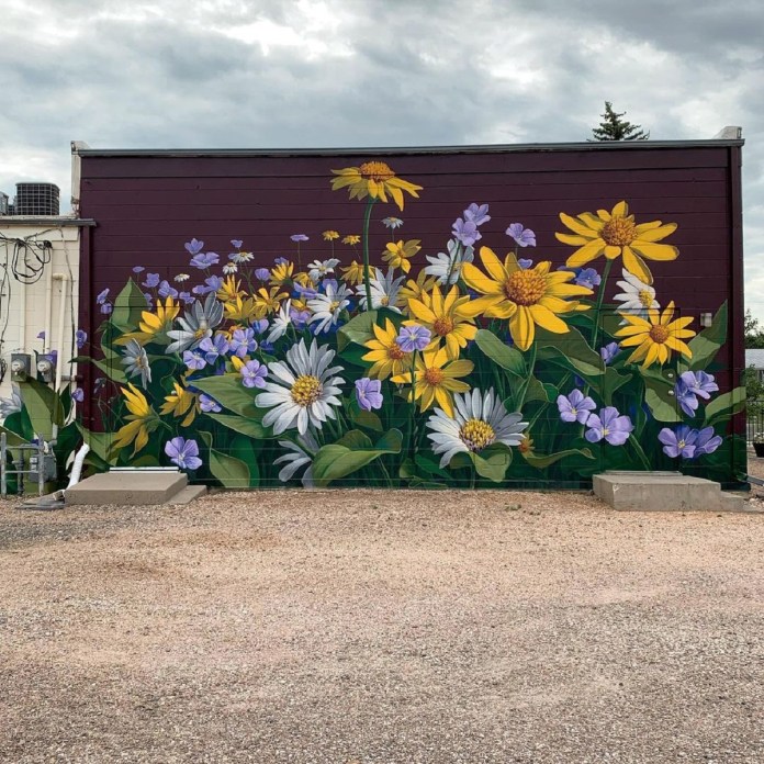 A mural of yellow sunflowers, white daisies, and purple wildflowers painted in a realistic style along a maroon wall in Nebraska, USA by street artist KOHIN.