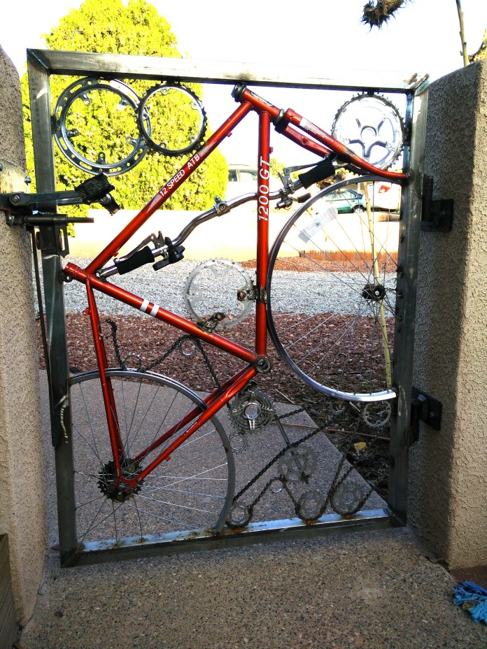 A red bicycle is welded into a rectangular metal gate, using its wheels and frame as structural elements. The gears and chains remain attached, and the gate opens between two concrete walls, with gravel and trees visible in the background.