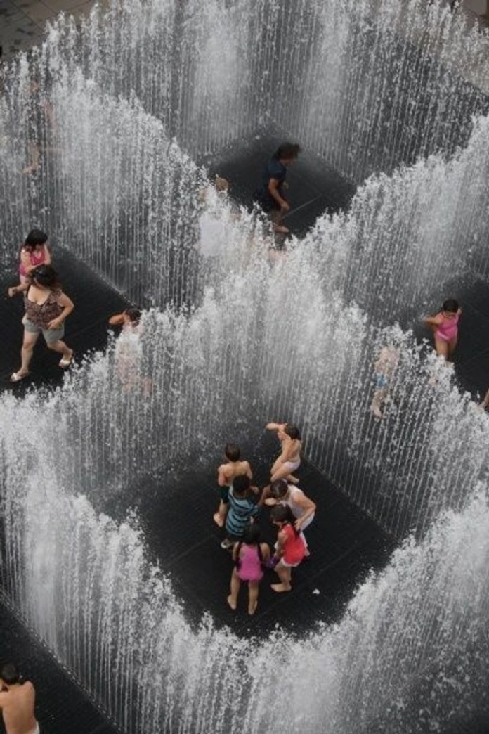 Overhead view of children playing inside a square maze created by vertical jets of water. The fountains form shifting walls, and the kids move in and out of the openings on a black ground grid.