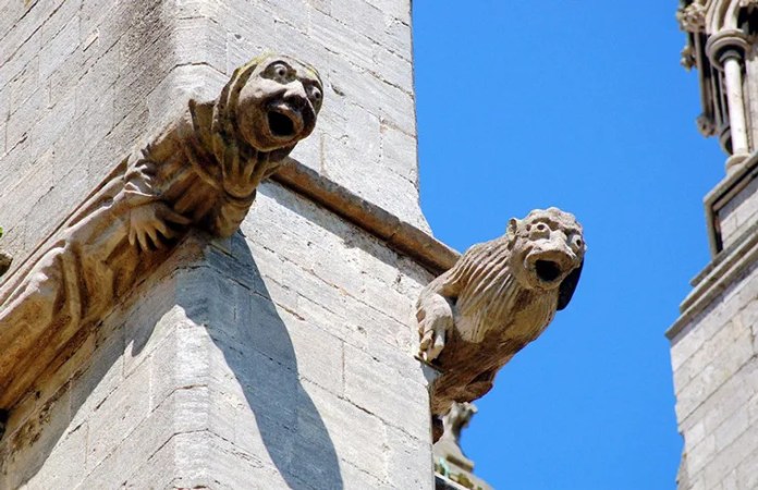 Two carved stone gargoyles with exaggerated, screaming faces protrude from the corner of a cathedral wall. Each has an open mouth and outstretched body, designed to channel rainwater away from the stone.