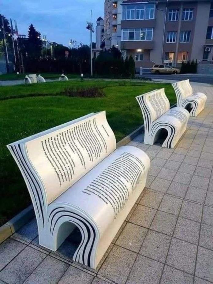 Curved white benches shaped like open books with black printed text across the surface. Installed along a paved path in a city park with grass and buildings in the background.