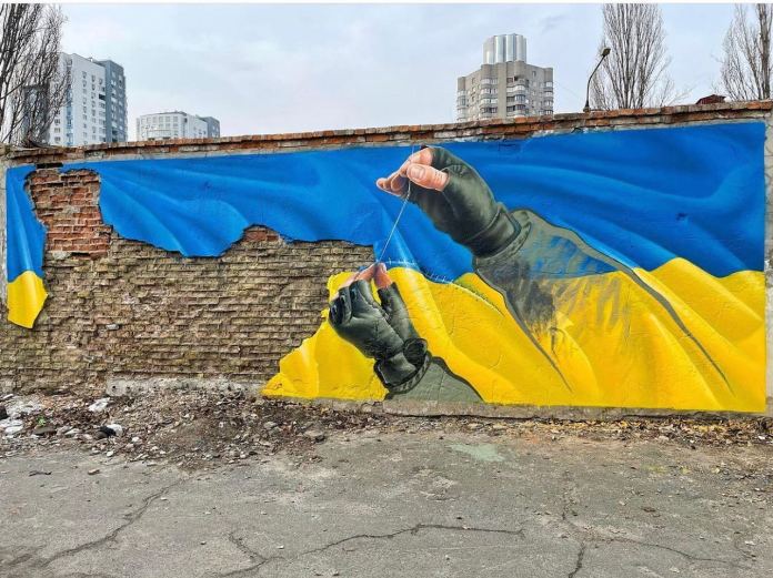 Mural by Sasha Korban in Kyiv, created during April 2022 amidst the battles for the liberation of Bucha and Irpin. The artwork depicts two gloved hands repairing a tear in the Ukrainian flag, symbolizing resilience and the nation's effort to heal amidst war. The vibrant blue and yellow of the flag contrast with the exposed brick wall, reflecting the scars of conflict. Korban's signature style, blending photorealism and Pop Art, highlights the theme of peace and recovery. The mural stands as a testament to Ukraine's enduring strength and the hope for a brighter future.