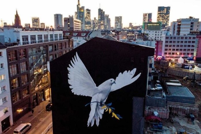Street art mural by German artist Justus Becker, located on the exterior of a downtown Frankfurt building. The artwork features a giant white dove of peace in mid-flight, holding an olive branch painted in the blue and yellow colors of the Ukrainian flag. The black background of the wall makes the dove stand out prominently, while the urban skyline of Frankfurt with modern buildings and warm evening lights adds context. Created over three days, the mural symbolizes hope, solidarity, and support for Ukraine during times of conflict.
