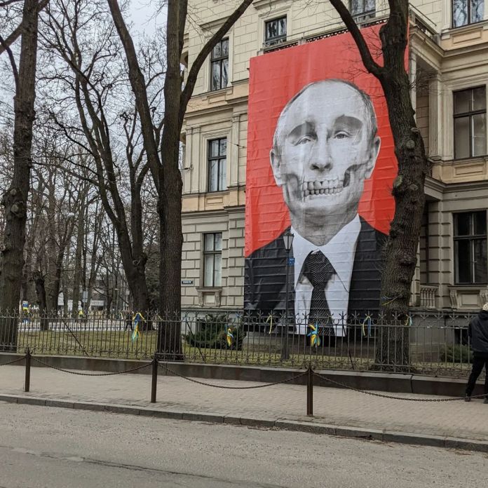 A powerful banner displayed on the facade of the Museum of the History of Medicine in Riga, Latvia, featuring a portrait of Vladimir Putin with his face morphed into a skull. The artwork is set against a striking red background and positioned directly across from the Russian Embassy, making a bold anti-war statement. Created by conceptual artist Krišs Salmanis, the banner was originally commissioned as the cover image for Ir magazine. Yellow and blue ribbons tied to the nearby fence emphasize solidarity with Ukraine, reinforcing the artwork's condemnation of the war.